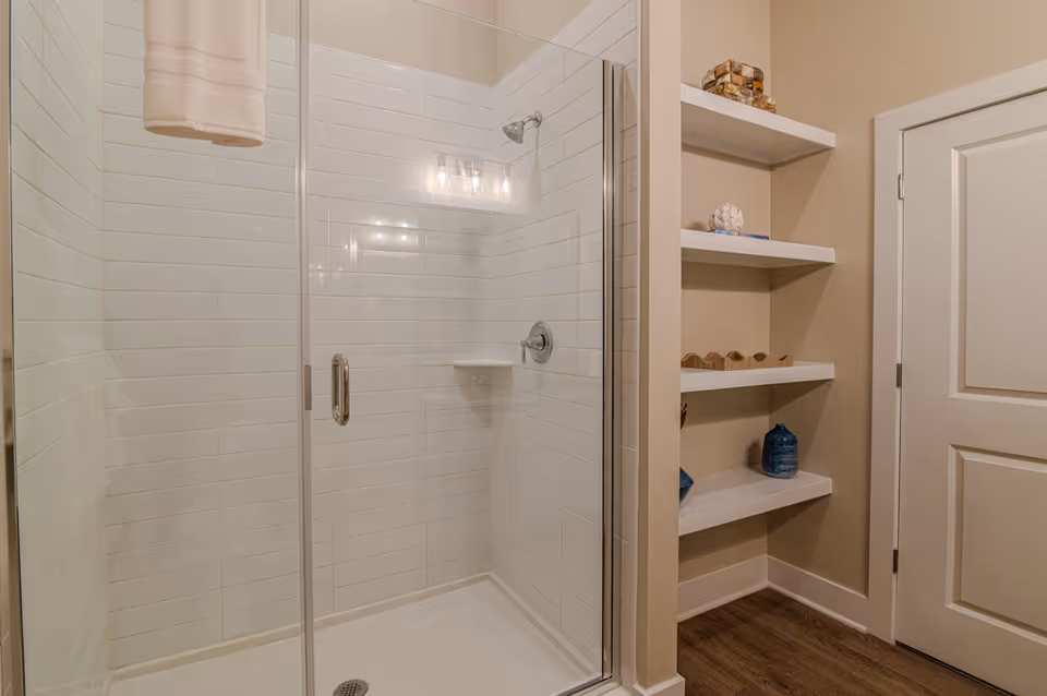 A clean, modern bathroom shower with white subway tiles and a glass door. To the right of the shower, there are built-in white shelves holding decorative items, and a closed white door is visible. The floor is wood-style laminate.