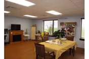 A cozy interior room with a wooden floor, a dining table covered with a yellow tablecloth set for four, a comfortable armchair near a window, a TV on a wooden stand, and a colorful painting on the wall.