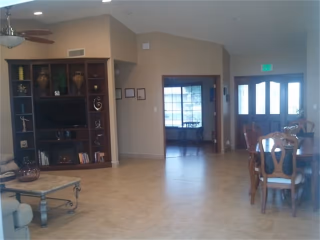 Interior view of a senior living facility showing a spacious room with a wooden dining table and chairs on the right, a built-in wooden entertainment center with a TV and decorative items on the left, a ceiling fan above, and a doorway leading to another room with large windows in the background.