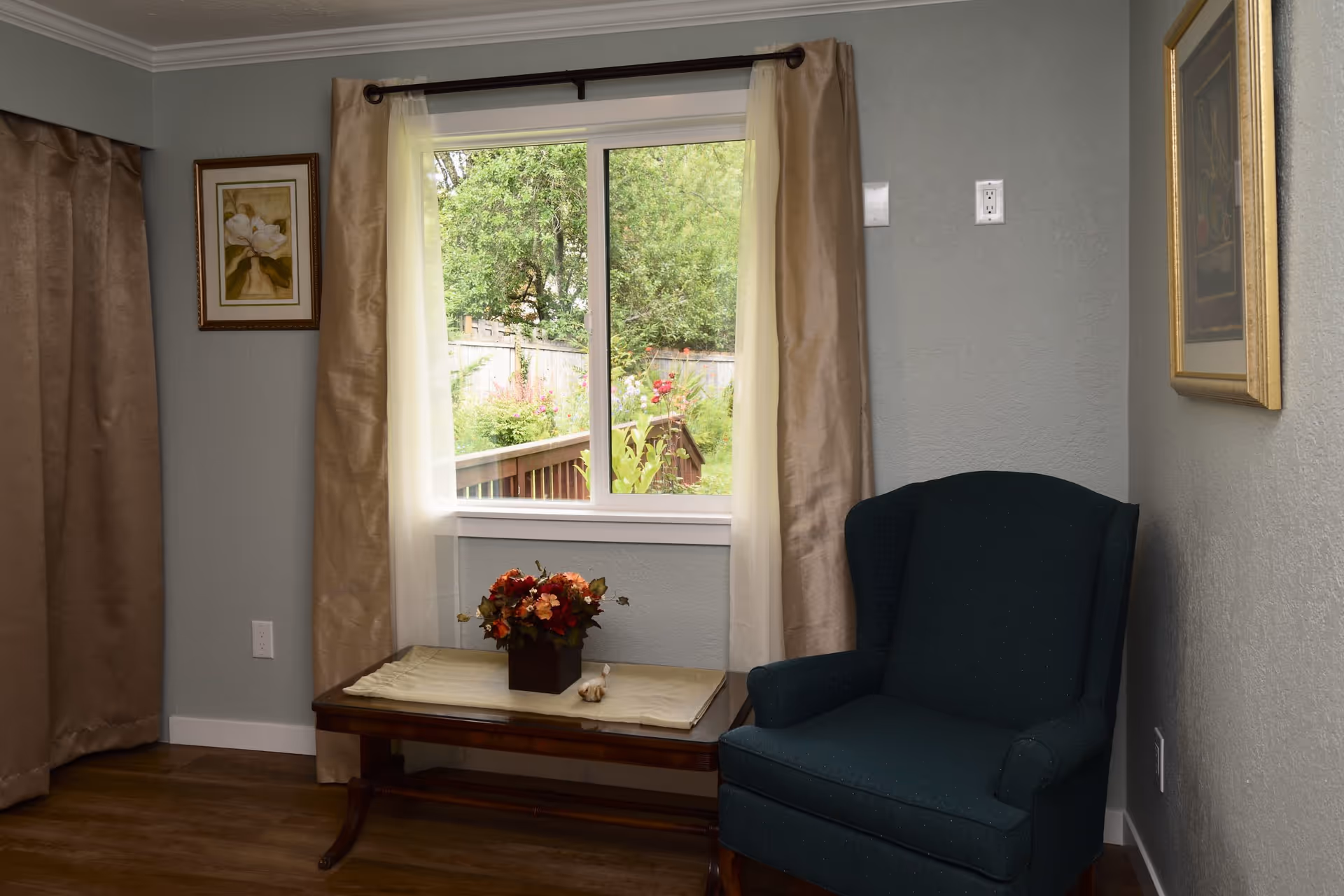 A cozy corner of a room with a dark upholstered armchair next to a wooden table holding a flower arrangement. A window with beige curtains shows a view of a garden with greenery and flowers outside. Two framed paintings hang on the light gray walls.
