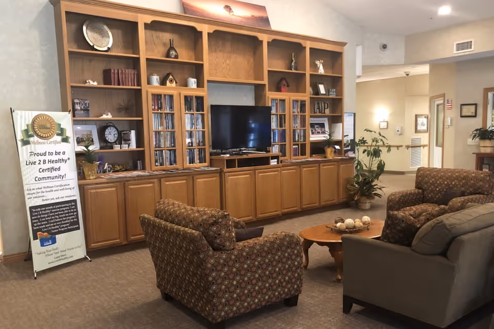 Lounge area with wooden built-in shelving and TV, upholstered armchairs and a coffee table in a senior living facility.