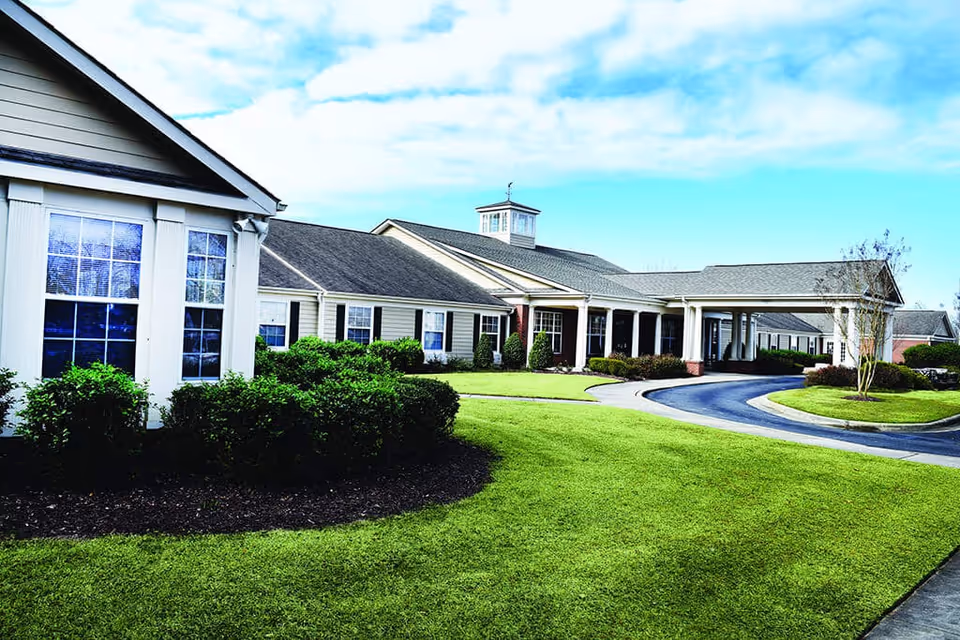 Exterior view of a single-story senior living facility building with beige siding, white trim, and multiple windows. The building has a covered entrance with columns and a circular driveway. The surrounding area features well-maintained green lawns, shrubs, and a partly cloudy sky.