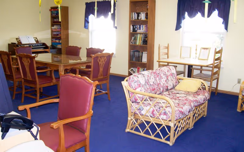 A communal lounge room with a floral wicker loveseat, upholstered chairs, a dining table, bookshelf and windows.