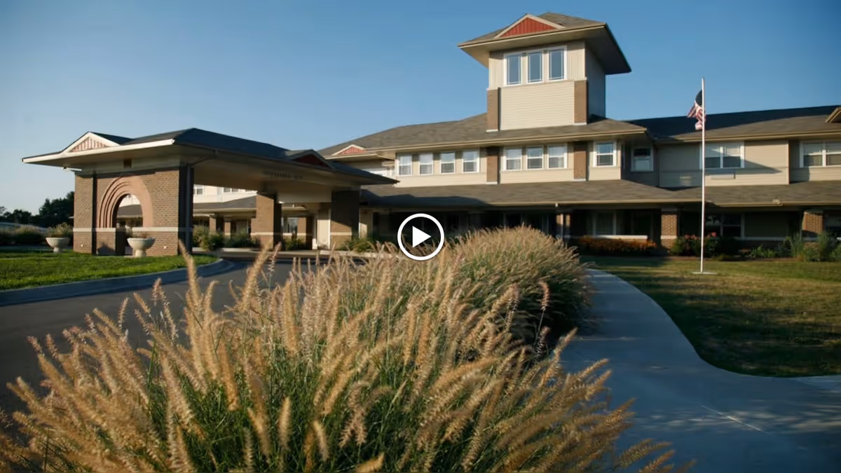 Exterior view of Prairie Living at Chautauqua, showing a large multi-story building with a covered entrance, surrounded by landscaped grass and ornamental grasses in the foreground under a clear blue sky.