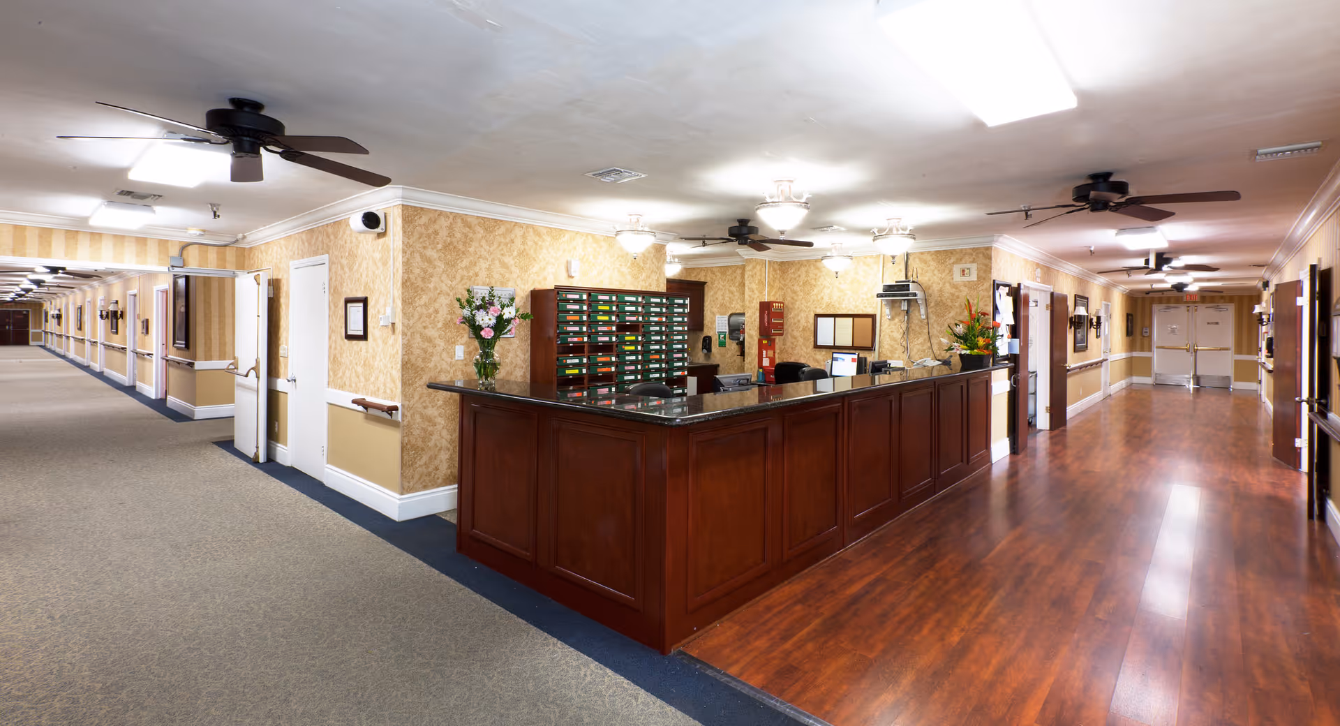 Interior view of a senior living facility hallway with a wooden reception desk in the center. The hallway has beige patterned wallpaper, ceiling fans, and overhead lights. There are doors along both sides of the hallway and a carpeted floor on one side and wooden flooring on the other. Flowers and office supplies are visible on the reception desk.