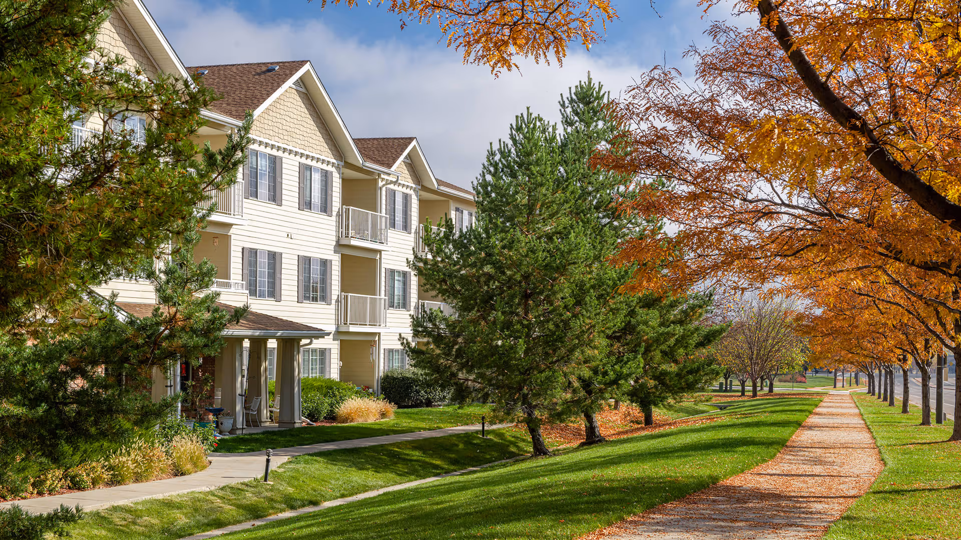 Exterior view of a multi-story residential building with beige siding and balconies, surrounded by green grass, trees with autumn foliage, and a paved walkway running alongside the building.