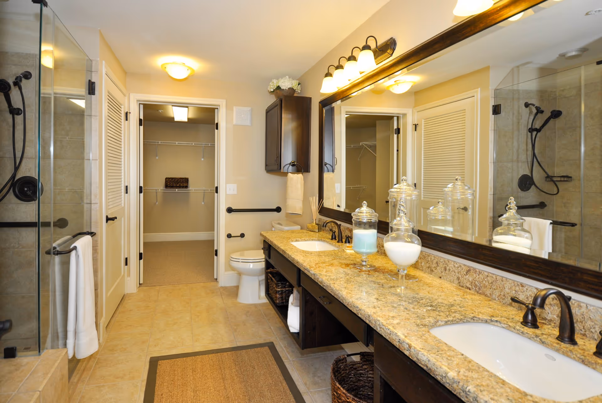 A spacious bathroom with a double sink granite countertop, large mirror, and dark wood cabinetry. There is a glass-enclosed shower with a handheld showerhead on the left. The bathroom has beige tiled floors and walls painted in a light cream color. A toilet is visible next to a doorway leading to a walk-in closet with wire shelving. The bathroom is well-lit with ceiling and vanity lights.