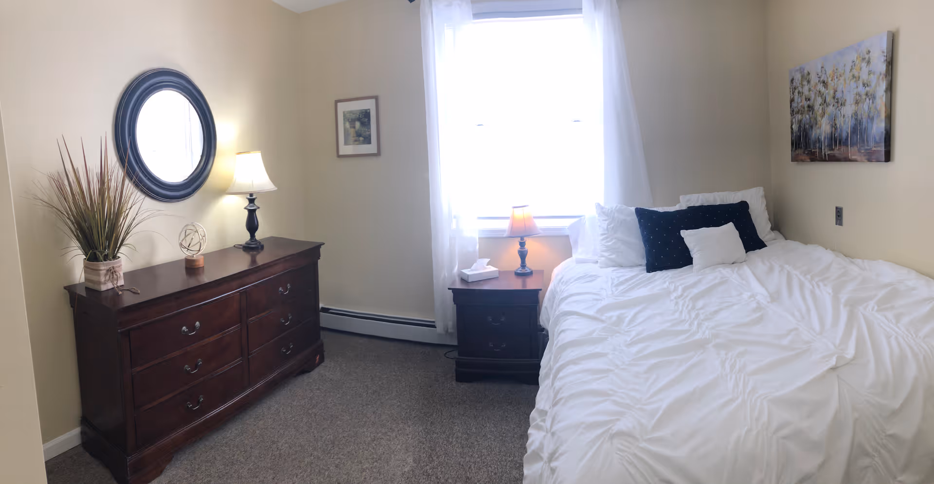 Bright bedroom with a neatly made bed, nightstand with a lamp, and a wooden dresser topped with a round mirror and decorative items.