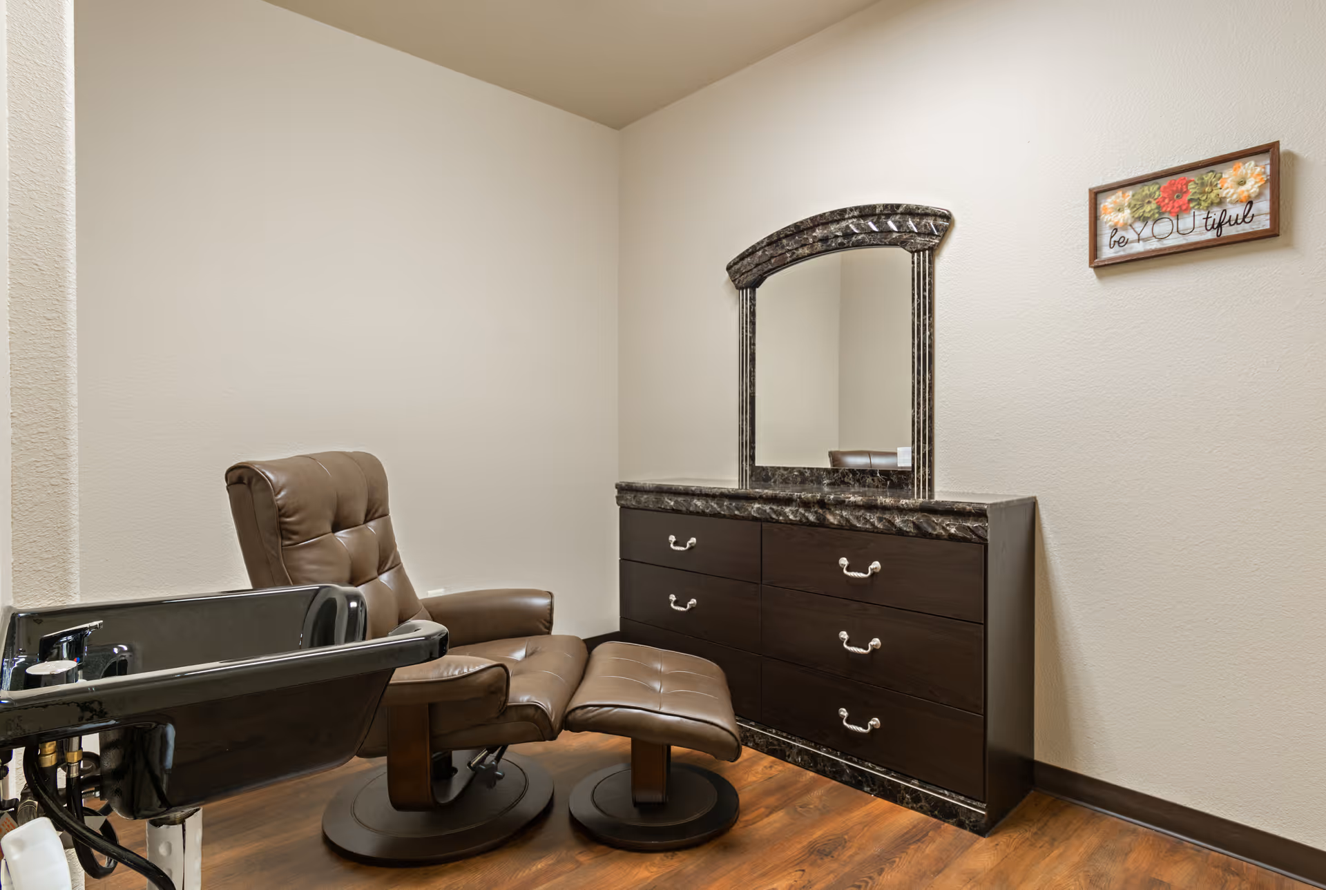 A small room with a brown leather reclining chair and matching footrest, a black wash basin for hair washing, and a dark wooden dresser with a large mirror. A decorative sign on the wall reads 'be YOU tiful' with colorful flowers.