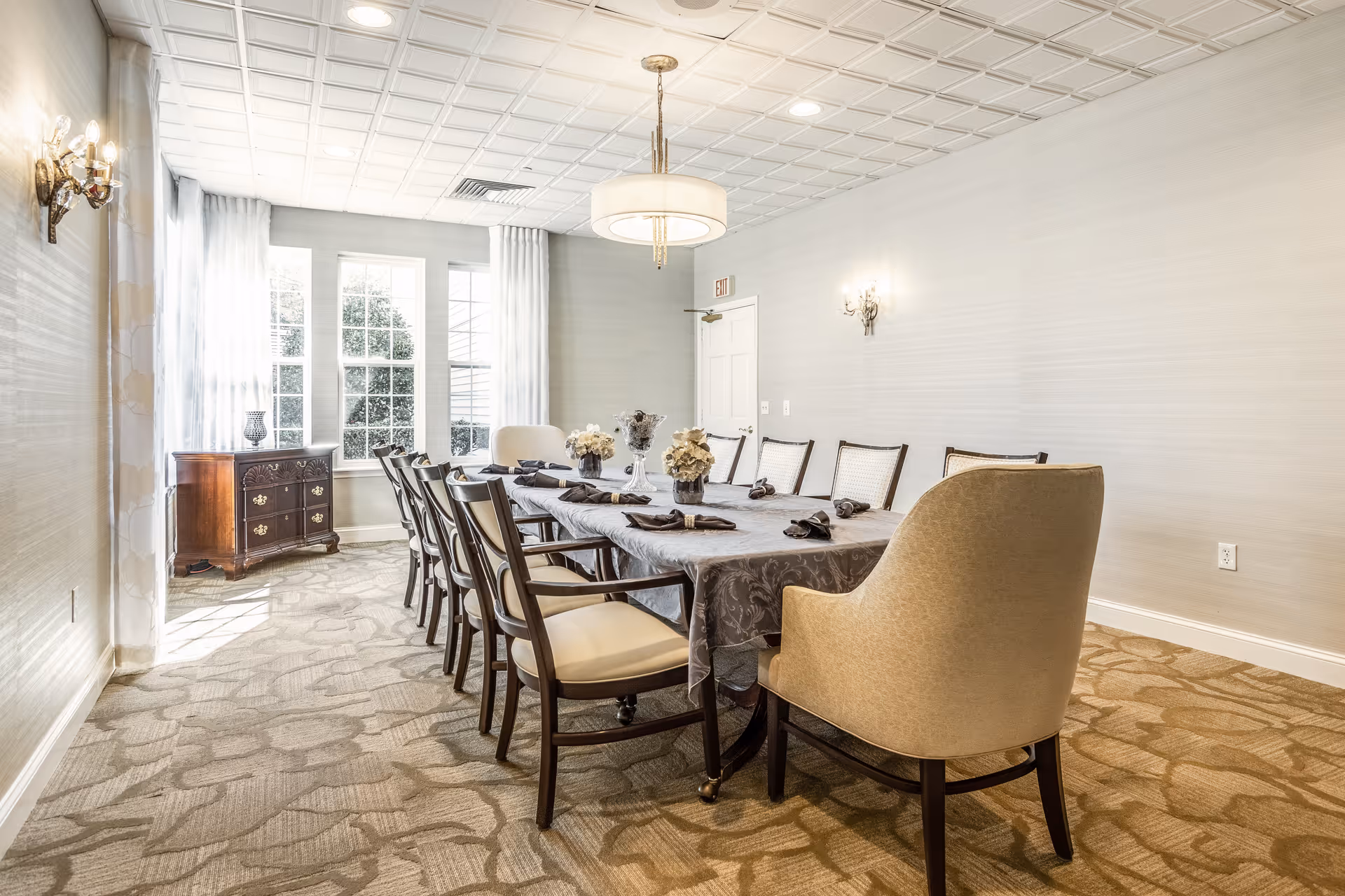 A bright dining room with a long rectangular table covered with a gray tablecloth, surrounded by ten chairs including two armchairs at the ends. The table is set with black napkins and decorative flower arrangements. The room has large windows with white curtains, beige patterned carpet, light gray walls, and a white ceiling with a modern chandelier and wall sconces providing lighting.
