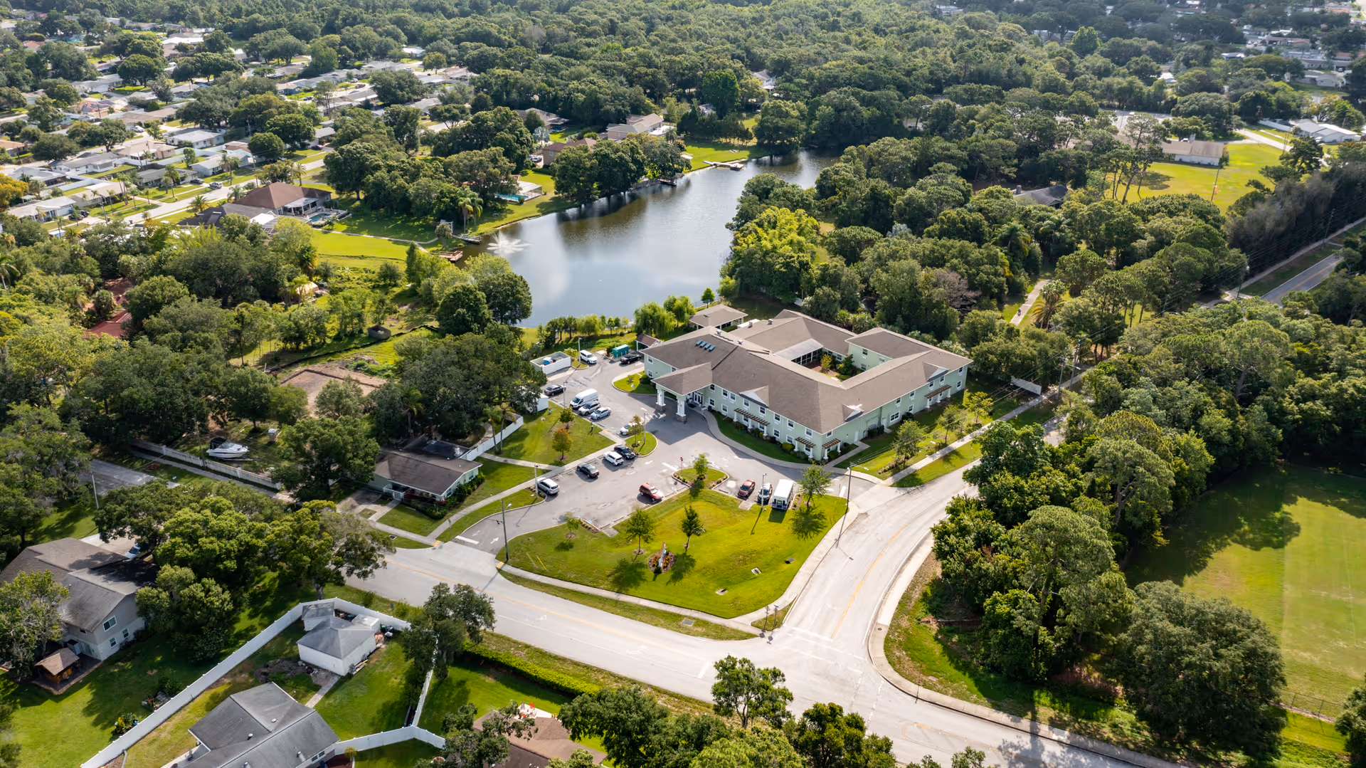 Aerial view of a senior living facility with a parking lot beside a small lake surrounded by trees and nearby houses.