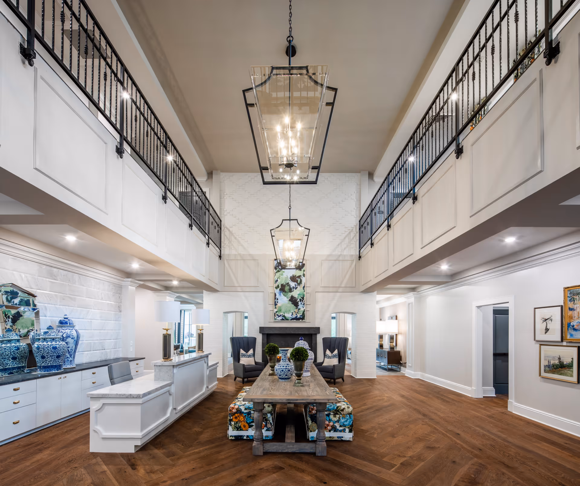 Spacious and elegant interior common area with high ceilings, two large hanging light fixtures, a wooden table with decorative vases and plants, floral upholstered benches, two armchairs near a fireplace, white paneled walls, and a balcony with black railings overlooking the room.