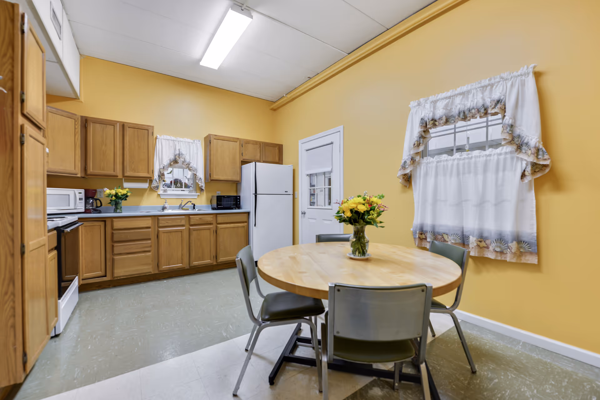 A kitchen with wooden cabinets, a white refrigerator, microwave, and stove. There is a round wooden table with four metal chairs around it. A vase with yellow flowers is placed on the table. The walls are painted yellow, and there are two windows with white curtains featuring a seashell pattern.