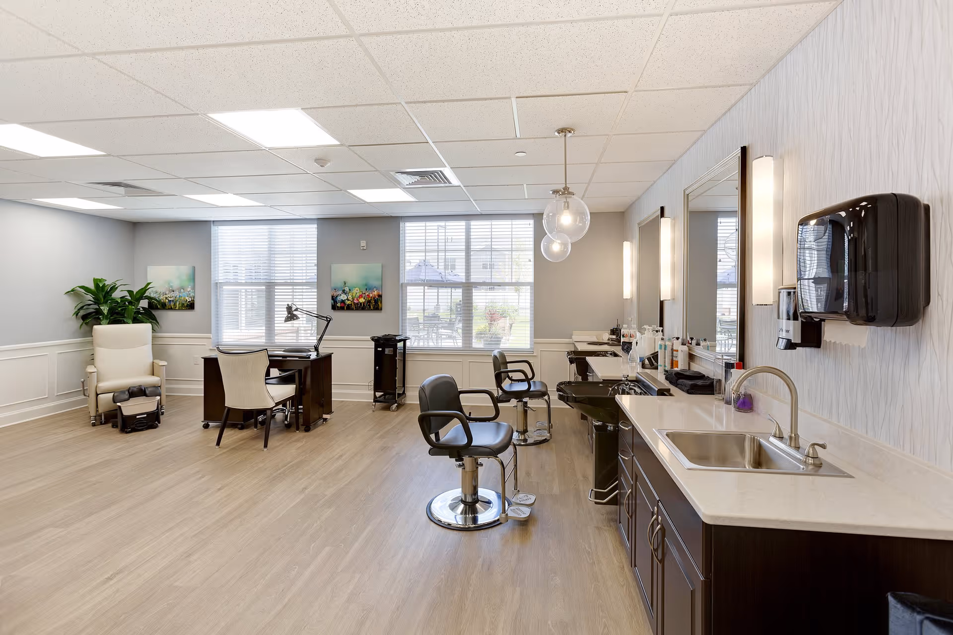 A bright and clean salon area in a senior living facility with two black salon chairs in front of mirrors and sinks, a desk with a chair and lamp near the window, a comfortable beige recliner chair with a footrest, and a plant in the corner. The room has large windows with blinds, light wood flooring, and modern lighting fixtures.