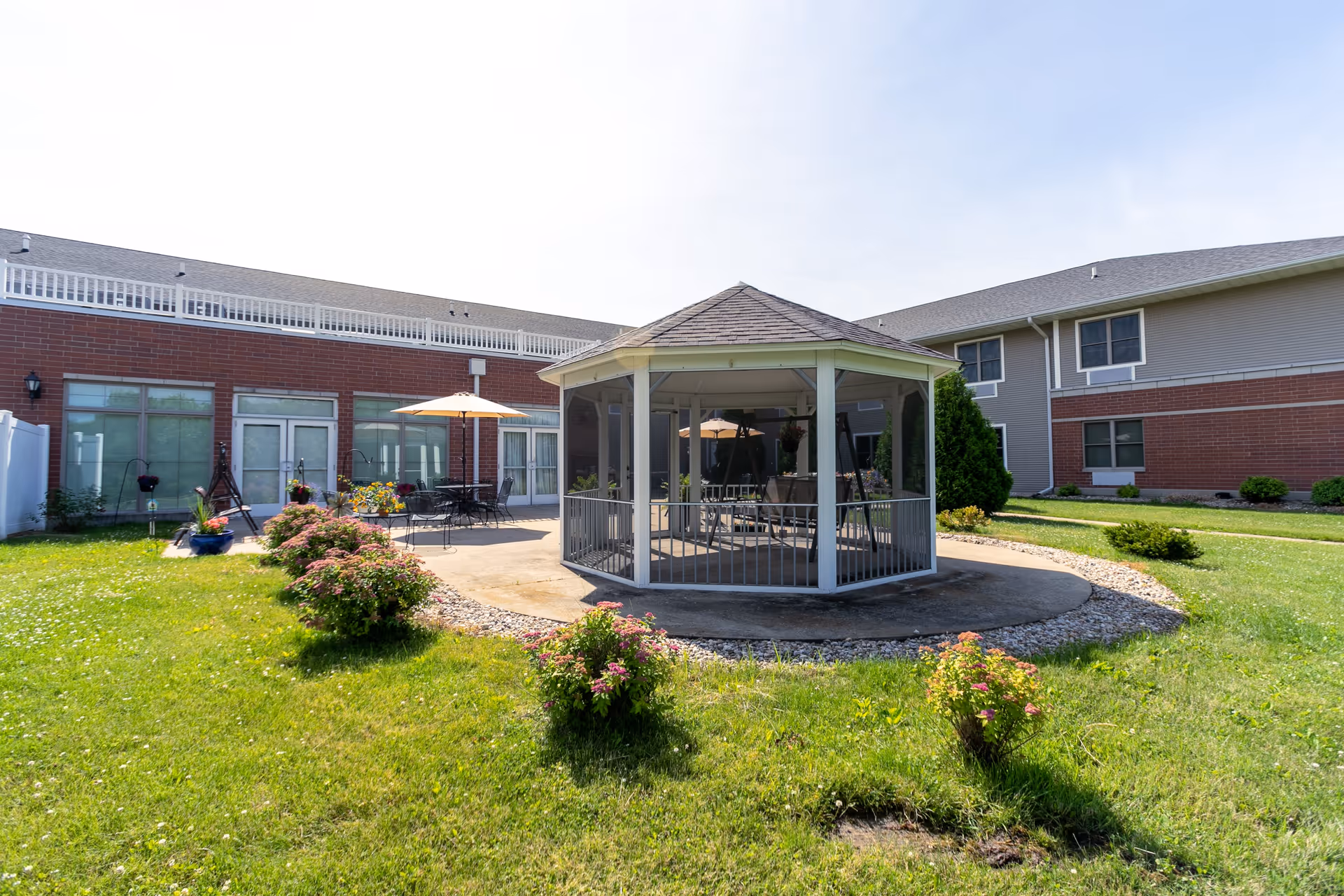 Outdoor courtyard area of a senior living facility featuring a central gazebo with seating, surrounded by green grass, small bushes, and a concrete patio with tables and umbrellas. The building with large windows and brick and siding exterior walls encloses the courtyard.