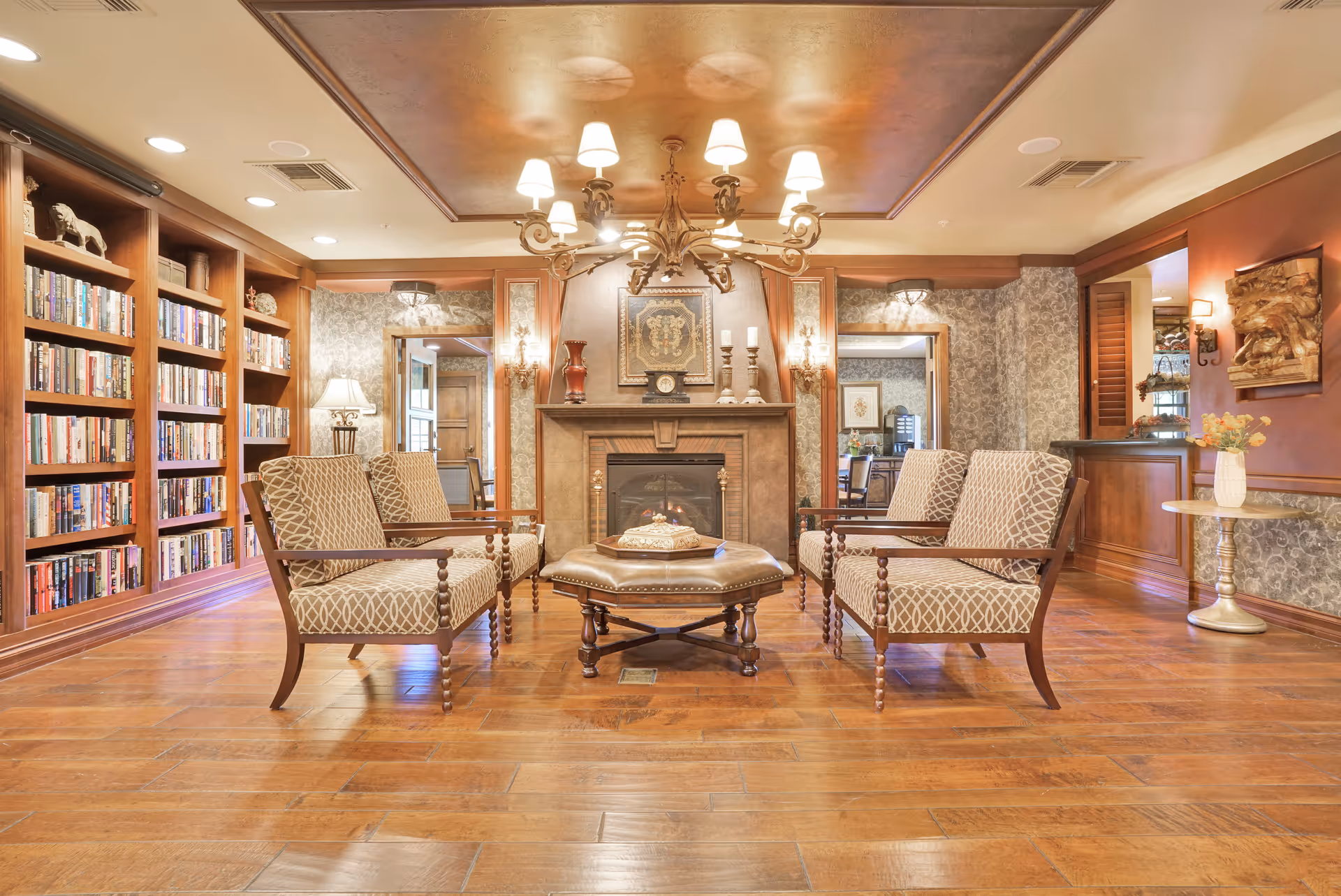 A cozy living room area with four patterned armchairs arranged around a wooden coffee table in front of a fireplace. The room features wooden flooring, built-in bookshelves filled with books on the left, ornate wallpaper, a chandelier hanging from the ceiling, and decorative items on the mantel and walls.
