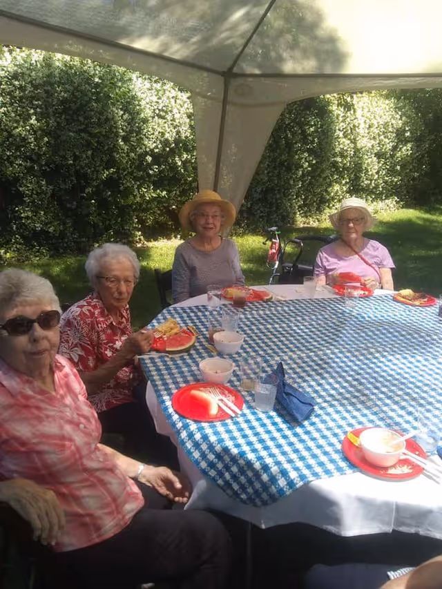 Four elderly women sitting around a table covered with a blue and white checkered tablecloth under a canopy outdoors. They are enjoying a meal with plates of food including watermelon and corn, with greenery in the background.