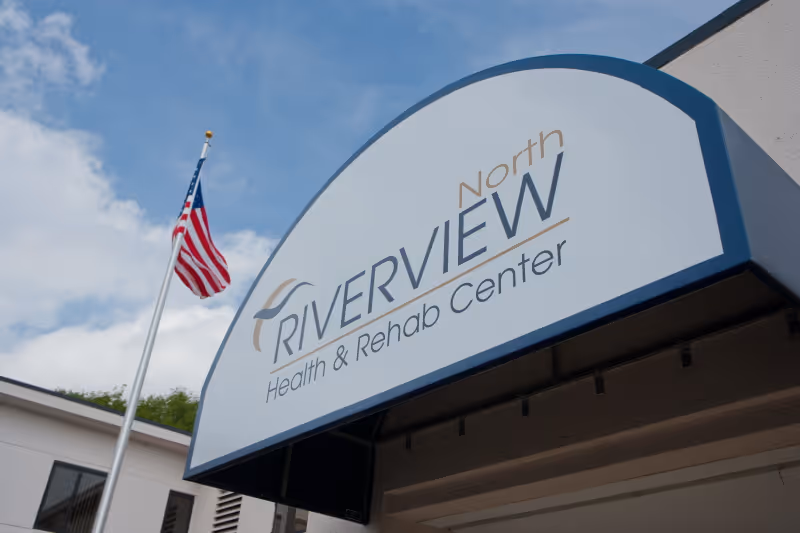 Entrance awning sign reading 'Riverview North Health & Rehab Center' beneath a blue sky with an American flag.