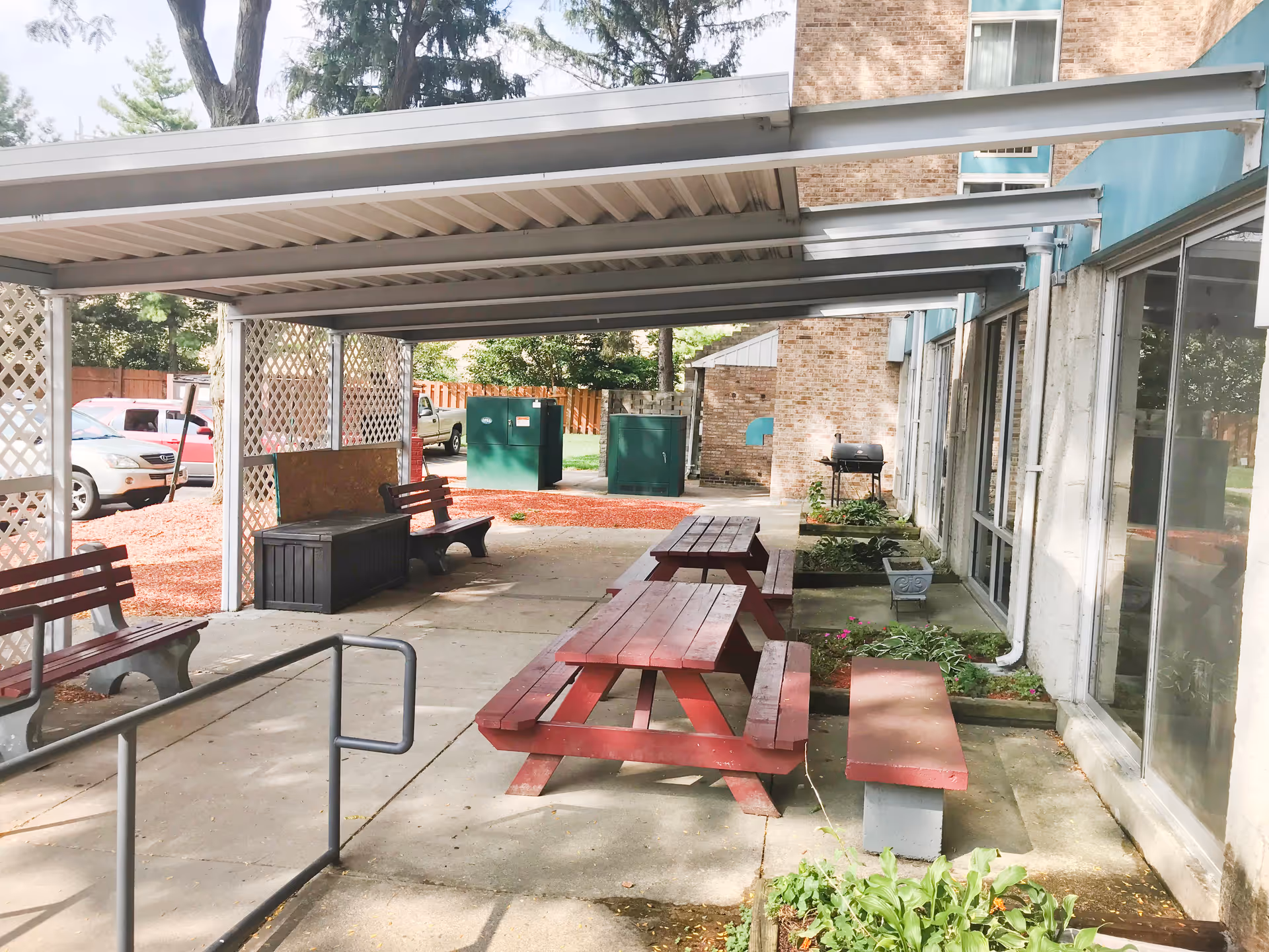 Outdoor covered patio area with red picnic tables and benches, a couple of wooden benches, a black storage box, and a barbecue grill near a brick building. There are some plants in small garden beds along the building and a parking area with cars in the background.