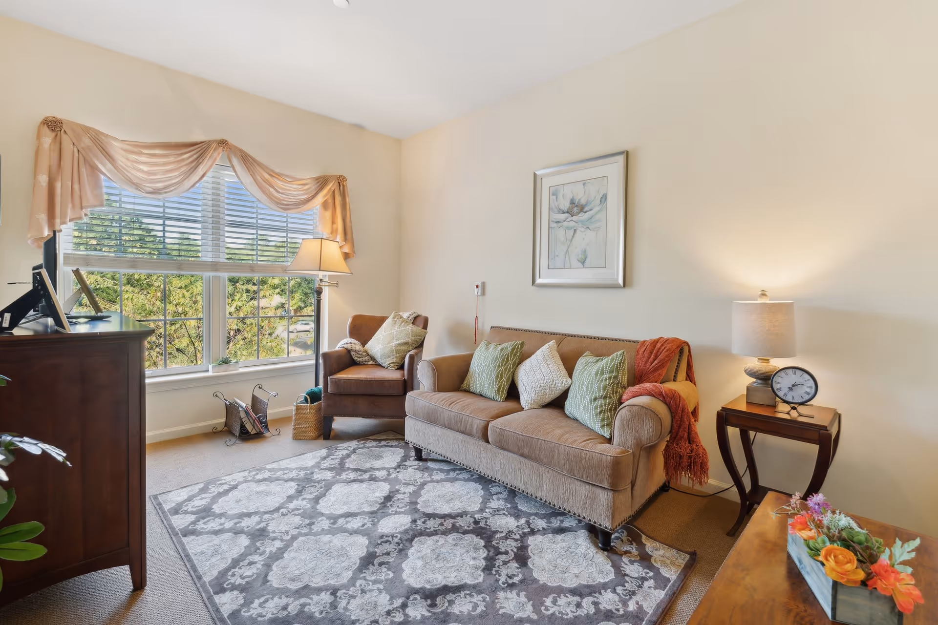 A cozy living room with a beige sofa adorned with green and white pillows and a red throw blanket. Next to the sofa is a wooden side table with a lamp and a clock. A brown armchair with a pillow is positioned near a large window with beige curtains, letting in natural light. A patterned area rug covers the floor, and a wooden cabinet with a TV is on the left side. A floral arrangement is on a wooden table in the foreground.