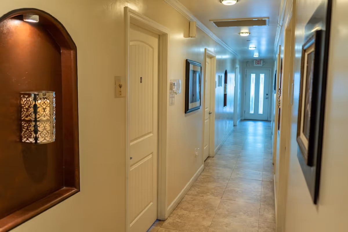 A well-lit hallway in a senior living facility with cream-colored walls and tiled floor. The hallway features several closed doors on both sides, framed pictures on the walls, and a decorative wall sconce on the left. At the end of the hallway, there is a door with glass panels and an exit sign above it.