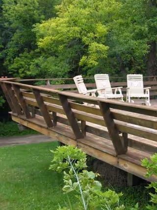 Wooden bridge with white chairs overlooks a green lawn and trees.