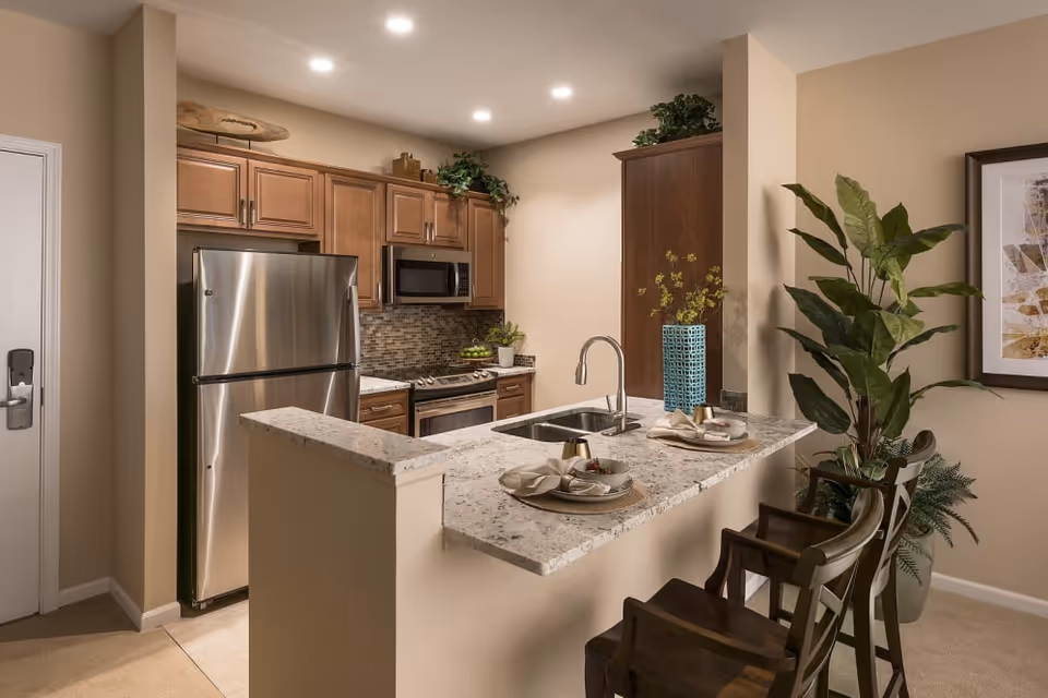 A modern kitchen area with a granite countertop island featuring a sink and two place settings with plates and napkins. The kitchen has wooden cabinets, a stainless steel refrigerator, microwave, and stove. There are decorative plants on top of the cabinets and a large potted plant next to the island. The walls are painted beige, and there is a framed artwork on the wall to the right.