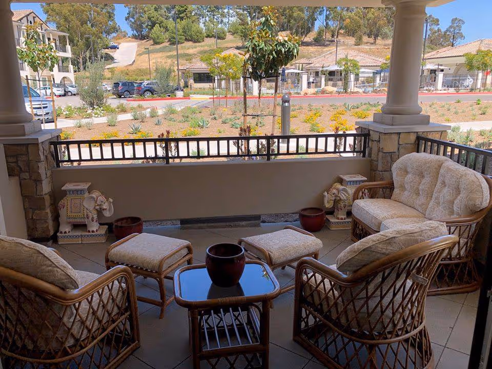 Covered outdoor patio area with cushioned wicker chairs, a loveseat, ottomans, and a small glass-top table. Two decorative elephant statues are placed near the corners. Beyond the patio, there is a landscaped garden with small plants and trees, a parking area with cars, and buildings in the background under a clear sky.