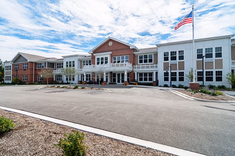 Front view of a two-story senior living facility with American flag and parking lot.