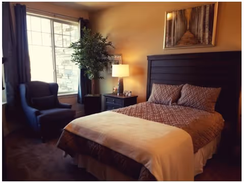 A cozy bedroom with a large window covered by dark curtains, a dark upholstered armchair near the window, a bed with a dark wooden headboard, brown quilted bedding, and a beige throw blanket. A nightstand with a lamp and framed photos is next to the bed, and a framed picture hangs on the wall above the headboard.