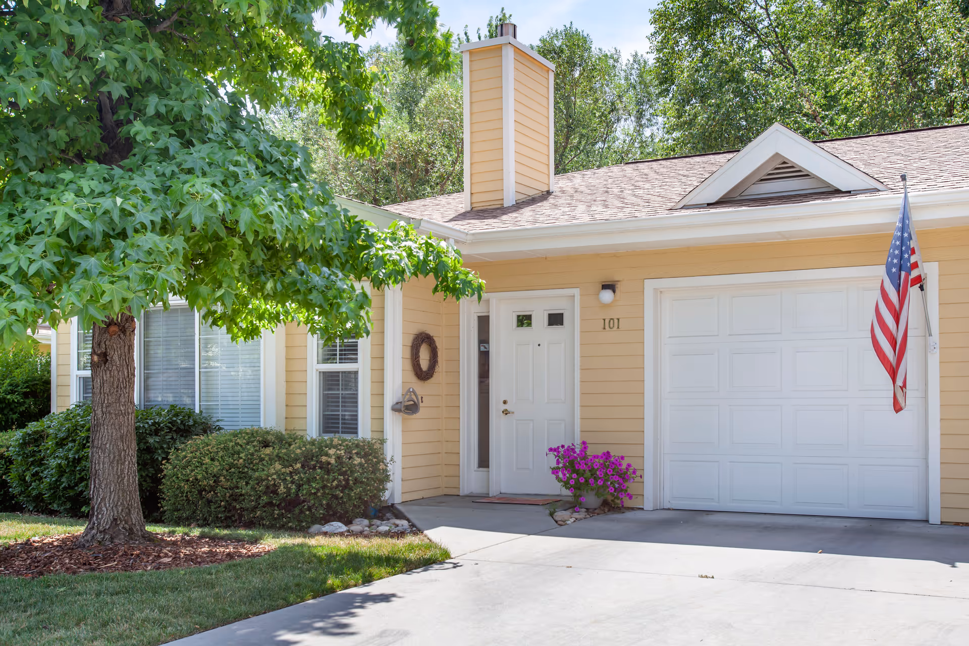 Front exterior of a yellow house with white door, garage, flag, and greenery.