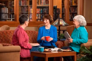 Three elderly women sitting on couches in a cozy living room with bookshelves in the background, engaging in conversation while holding a tablet, a book, and a red mug.