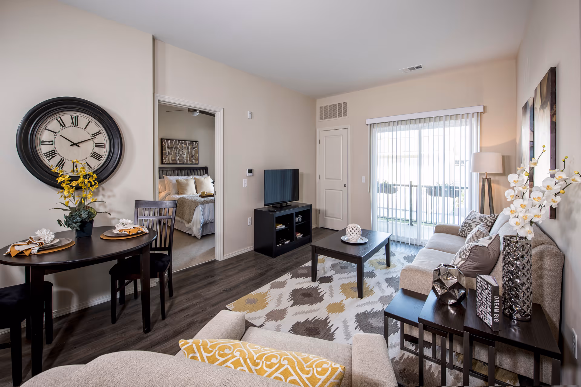A modern living room with a beige sofa adorned with patterned pillows, a dark wood coffee table on a patterned rug, and a TV on a black stand. To the left, there is a small dining area with a round table set for two and a large wall clock above it. A doorway leads to a bedroom with a bed and decorative pillows. Sliding glass doors with vertical blinds allow natural light into the room. The walls are painted light beige, and there are decorative flowers and artwork in the room.