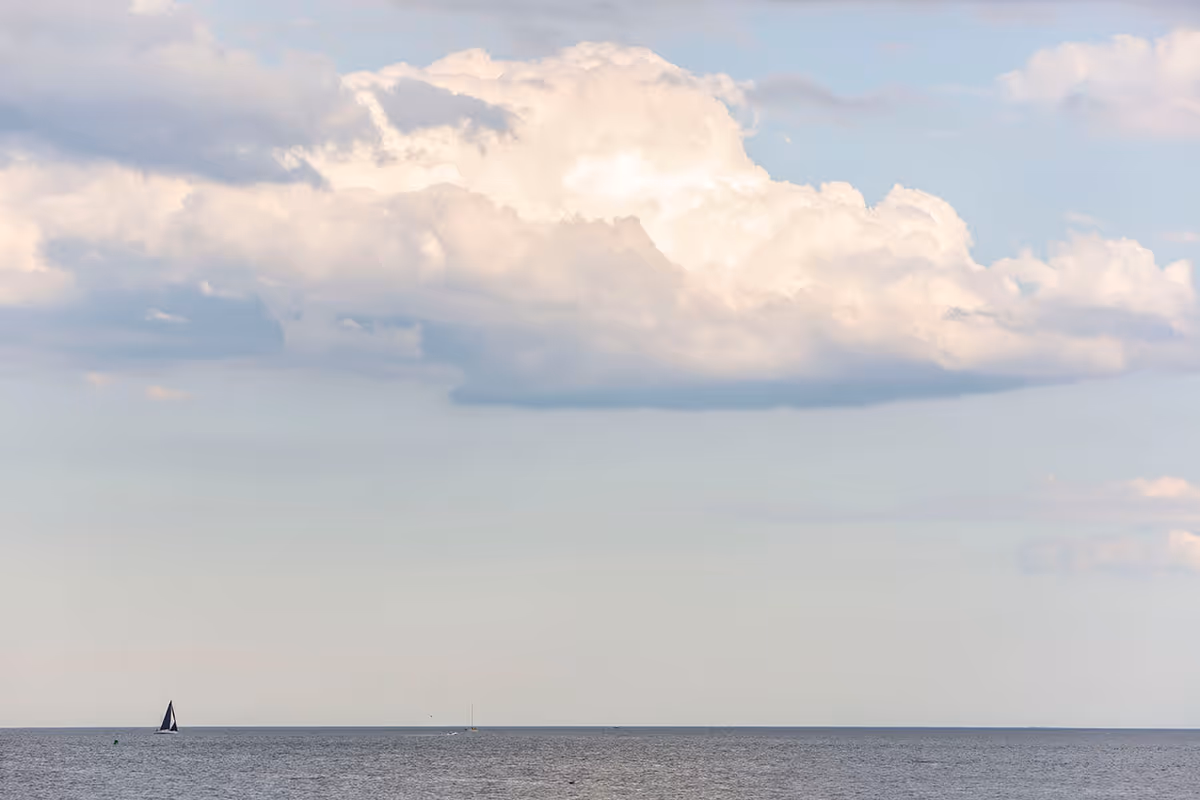 A calm ocean scene with a small sailboat on the water under a partly cloudy sky with large, fluffy clouds.