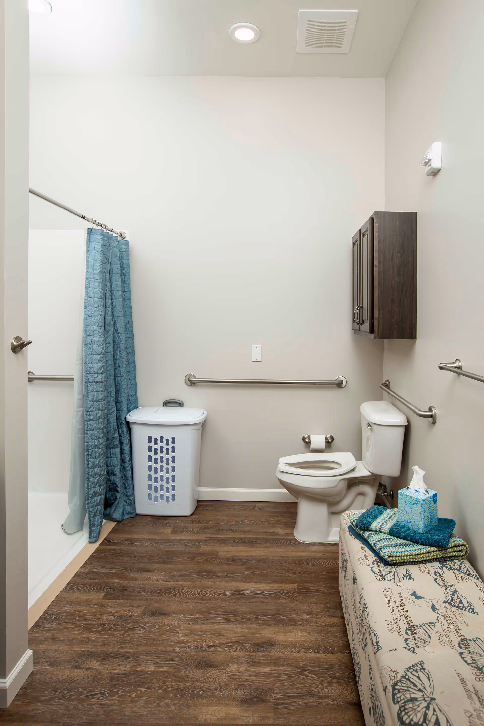 Accessible bathroom with a toilet, grab bars, a shower with a blue curtain, a laundry basket, wall cabinet, and a bench with folded towels and tissues.