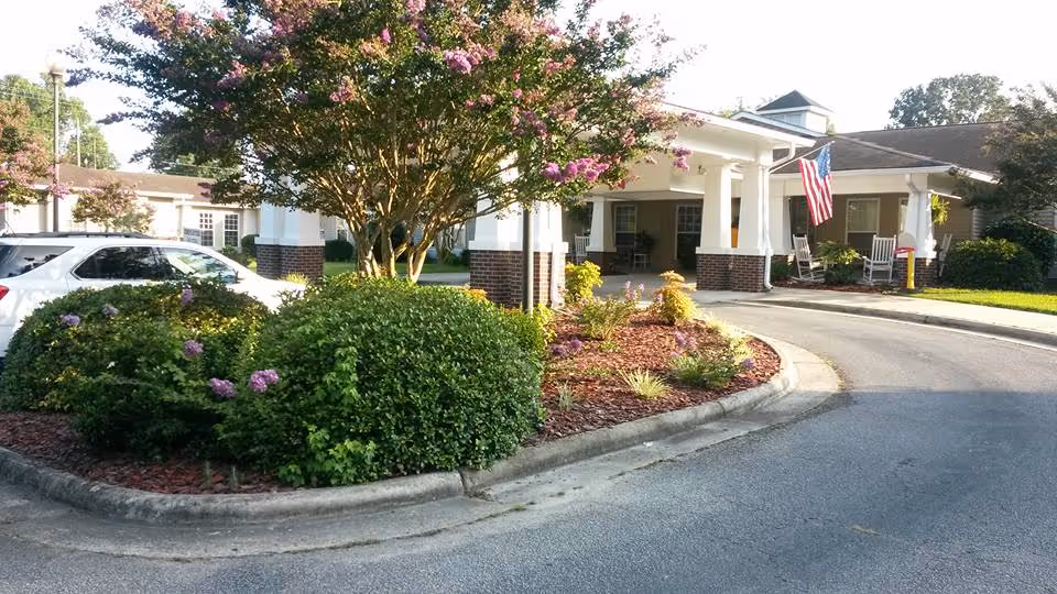 Entrance area of Somerset Court of Rocky Mount featuring a curved driveway, landscaped garden with bushes and flowering trees, a white car parked on the left, and a building with a covered porch displaying an American flag and rocking chairs.