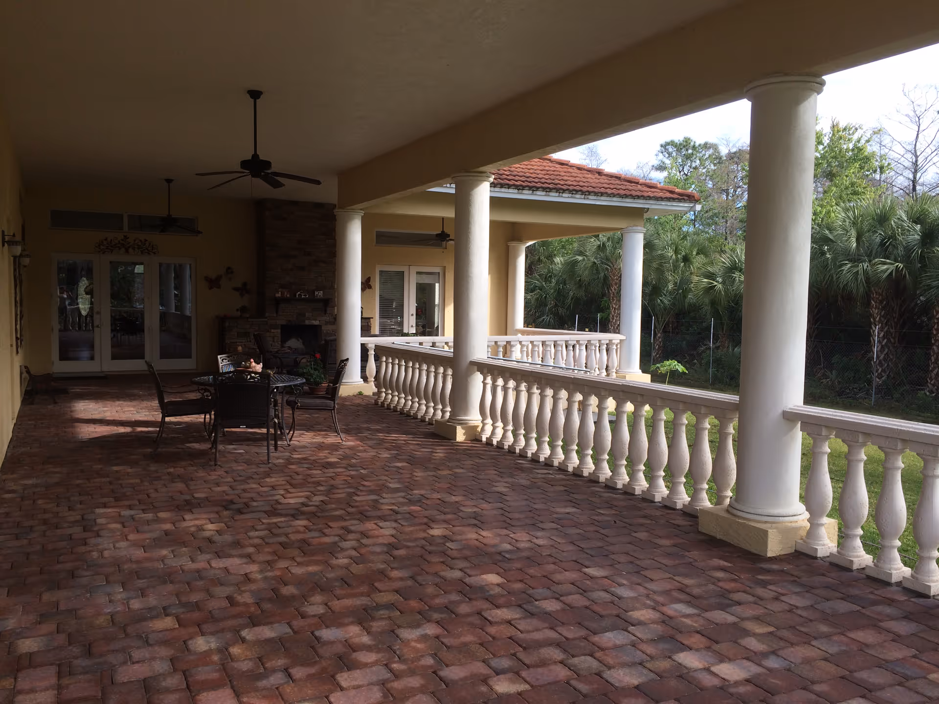 Covered outdoor patio area with brick flooring, white columns, and a white balustrade railing. There is a black metal table with four chairs in the middle, ceiling fans above, and a stone fireplace against the wall near glass doors. The patio overlooks greenery with palm trees in the background.