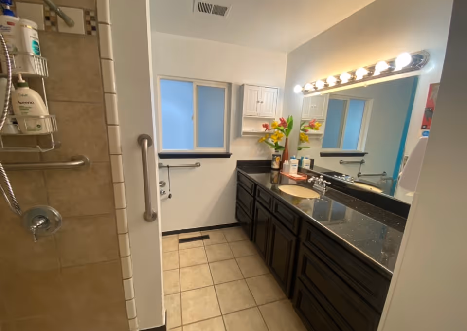 A bathroom with a tiled shower area on the left featuring a handheld showerhead and several bottles of toiletries on a hanging rack. The room has a large mirror above a black granite countertop with a sink and dark wooden cabinets below. There is a window with frosted glass on the far wall, a towel bar, and a grab bar near the shower entrance. The floor is tiled, and the lighting above the mirror consists of multiple round bulbs.