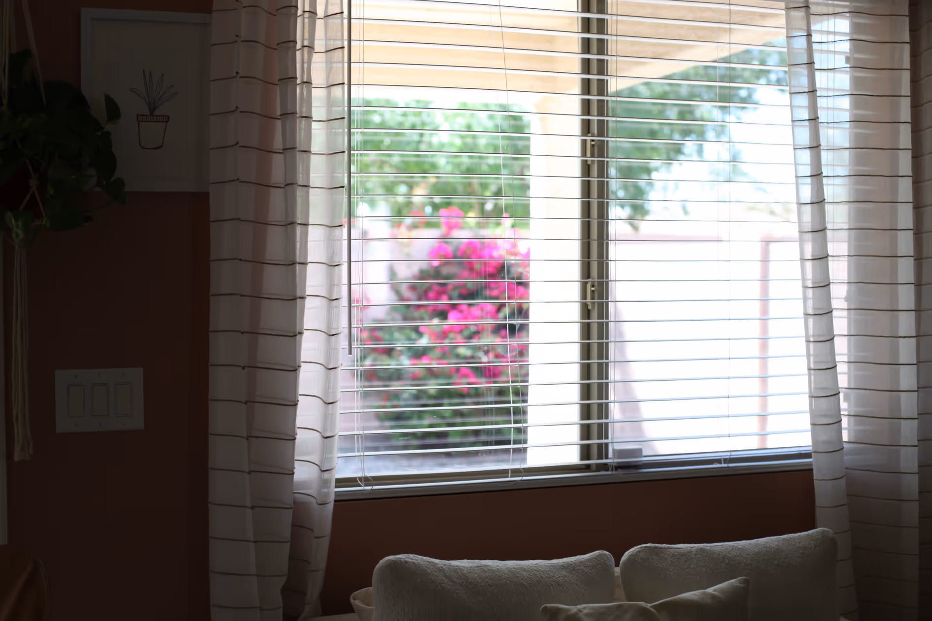A window with white horizontal blinds partially open, sheer white curtains with brown stripes on either side, and a view of pink flowers and greenery outside. Inside, there is a white cushioned seat or sofa below the window, and a wall with a light switch and a framed picture of a potted plant.