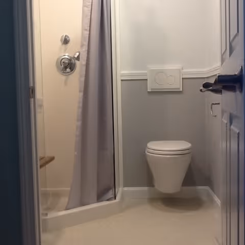 Small bathroom with a wall-mounted toilet and a shower area with a curtain. The walls are painted light gray and white, and the floor is a light beige color. A door with a handle is partially open, leading into the bathroom.