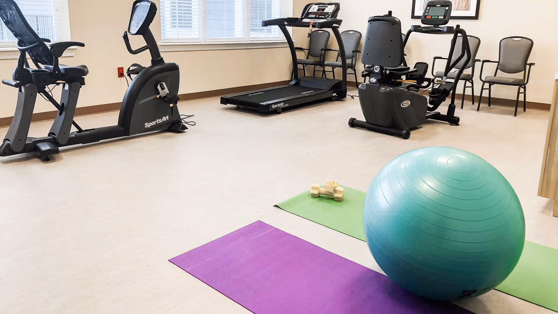 Exercise room with fitness equipment including a recumbent bike, treadmill, and an upright bike. There are several chairs lined up against the wall, two exercise mats on the floor with a pair of dumbbells on one mat, and a large blue exercise ball on the other mat. The room has large windows with white blinds.