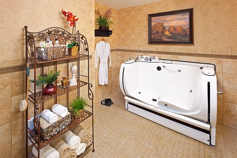 A well-decorated bathroom featuring a large white walk-in bathtub with control knobs, a white bathrobe hanging on the wall, a pair of black slippers on the floor, a metal shelving unit holding rolled towels, plants, and bath items, beige tiled walls and floor, and a framed landscape painting on the wall.