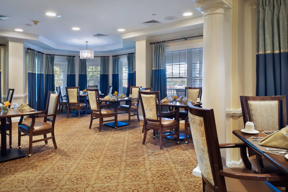 Elegant dining room with tables set for service, upholstered wooden chairs, patterned carpet, and large windows with blue drapes.