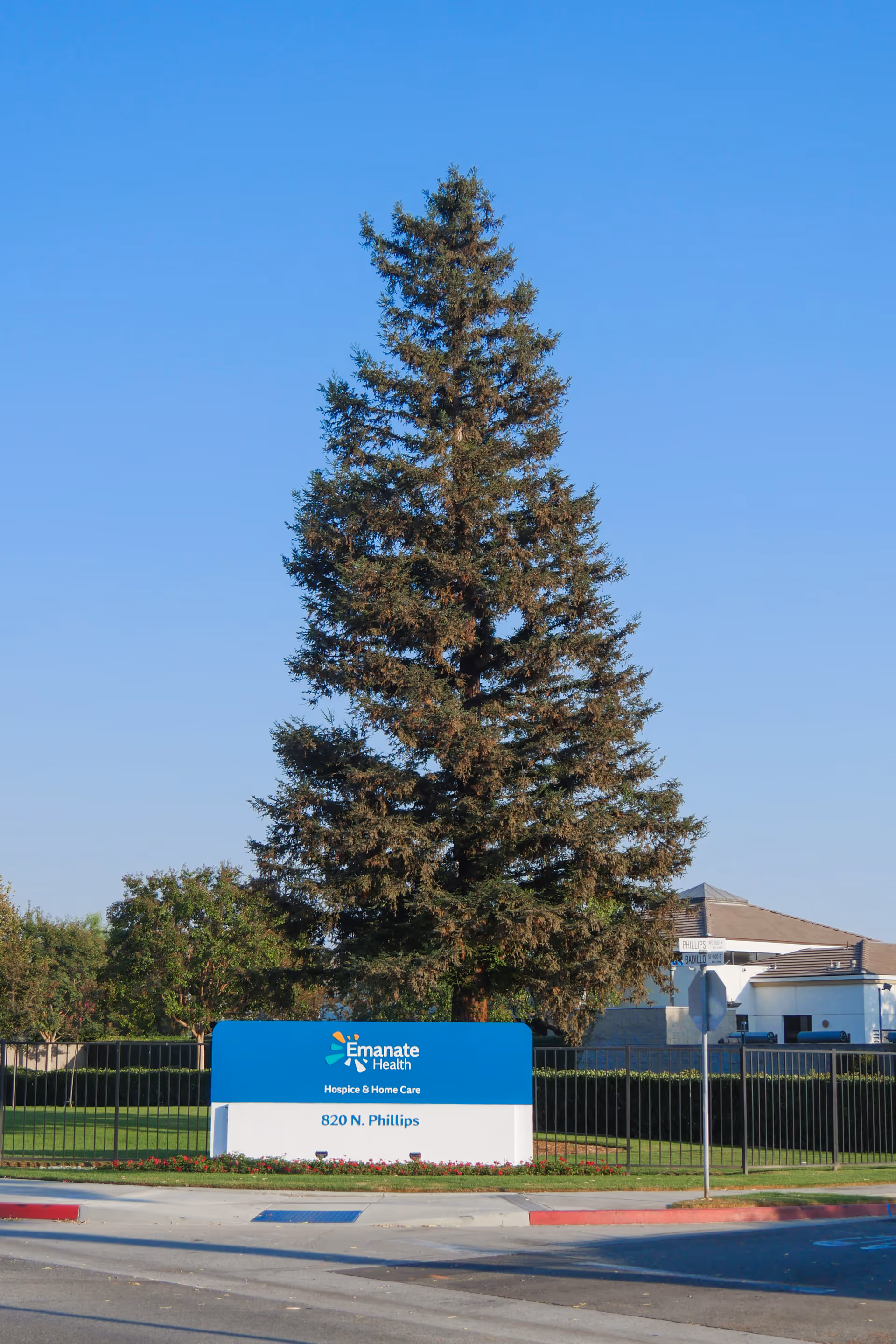 Outdoor view of Emanate Health Hospice & Home Care facility sign at 820 N. Phillips, with a large tree behind the sign and a building in the background under a clear blue sky.
