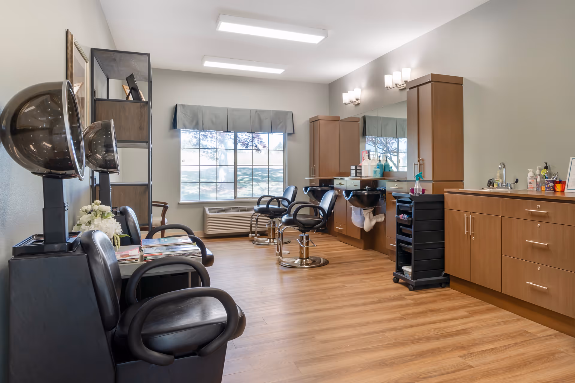 Interior salon room with hair styling chairs, hooded dryers, shampoo sinks and wooden cabinetry under ceiling lights and a large window.