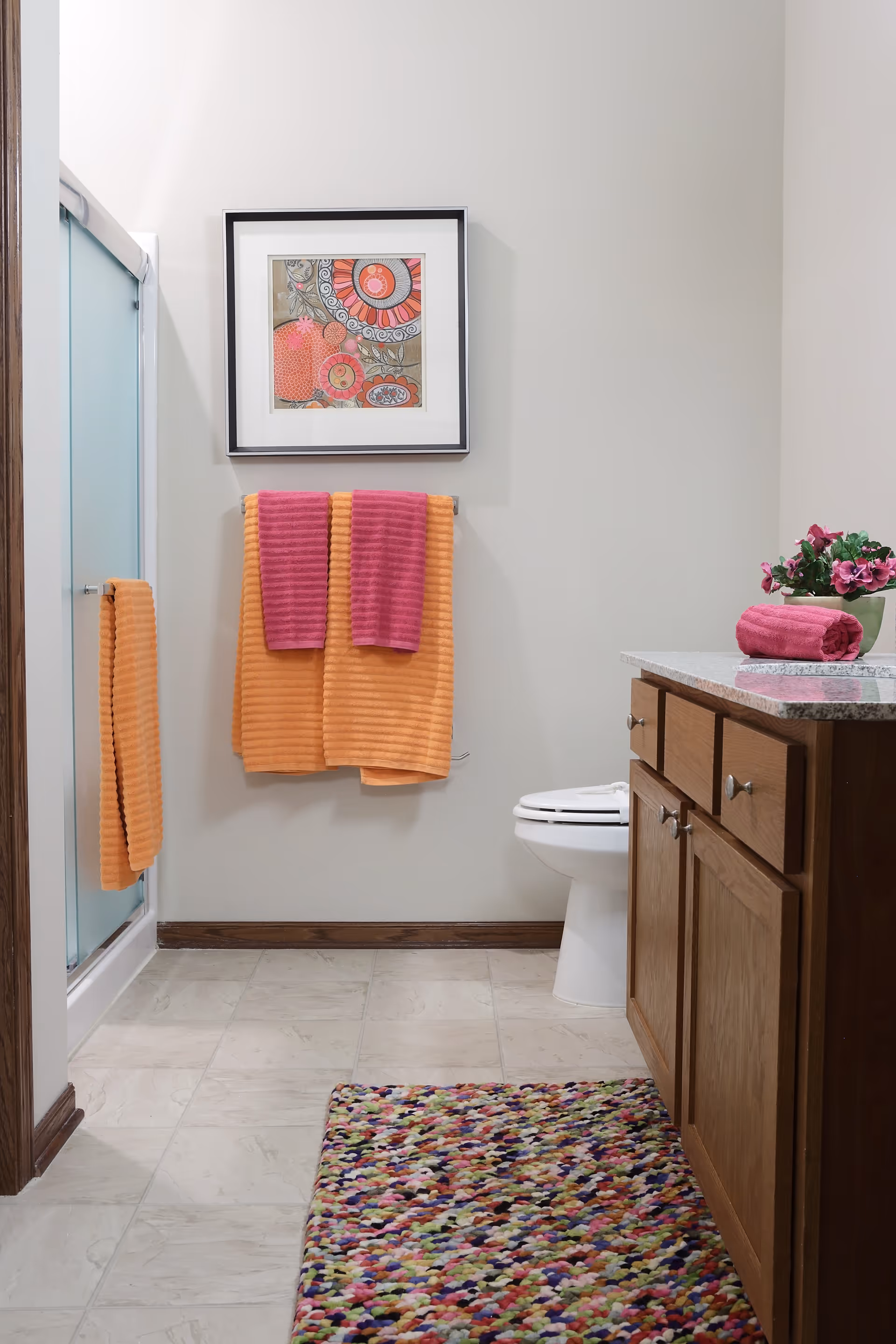 Bright bathroom with a wooden vanity, toilet, glass shower, colorful towels, framed artwork and a multicolored rug.