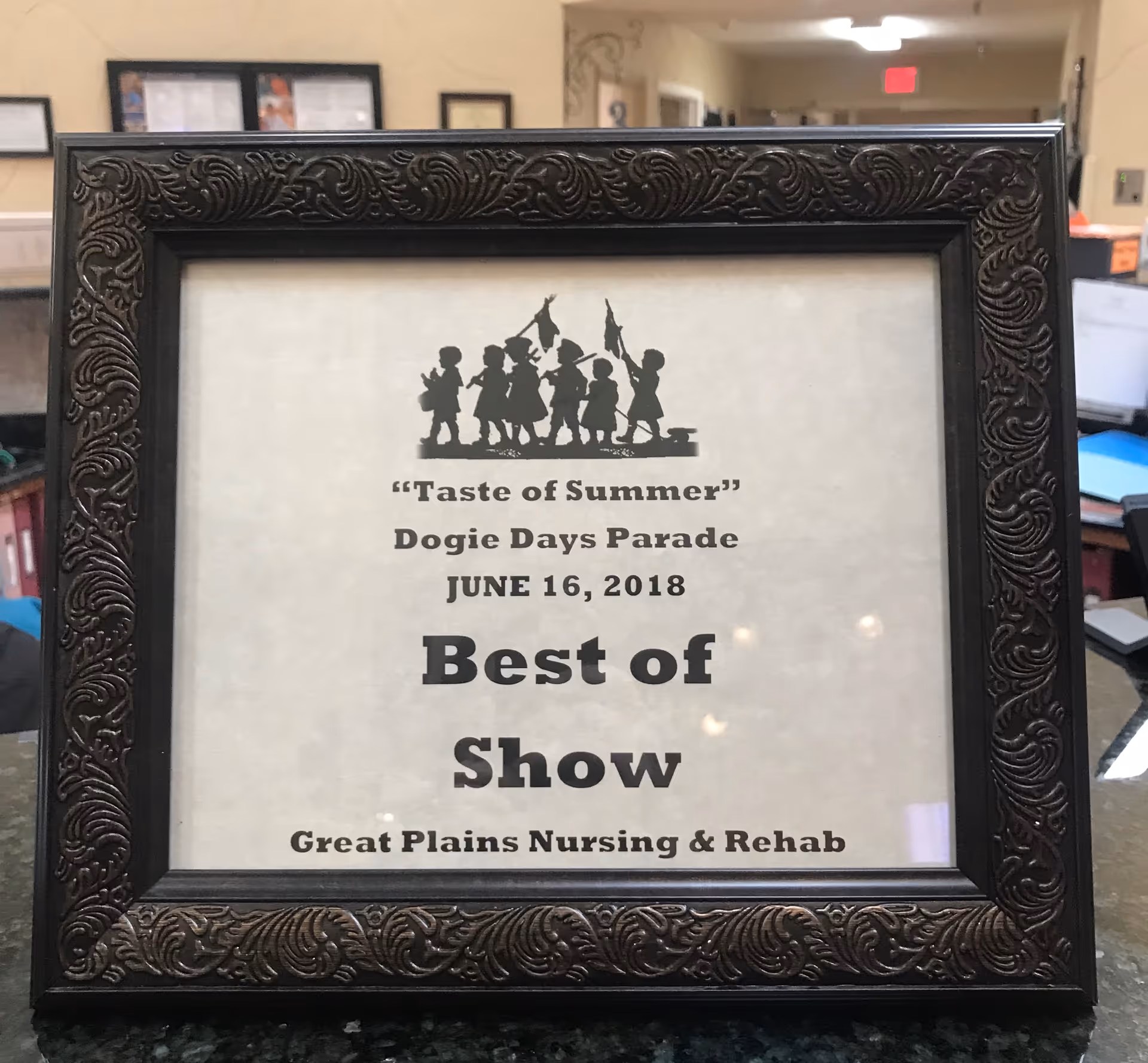 A framed certificate with an ornate dark frame on a countertop. The certificate reads: 'Taste of Summer' Dogie Days Parade June 16, 2018 Best of Show Great Plains Nursing & Rehab. The certificate features a silhouette of children holding flags at the top.