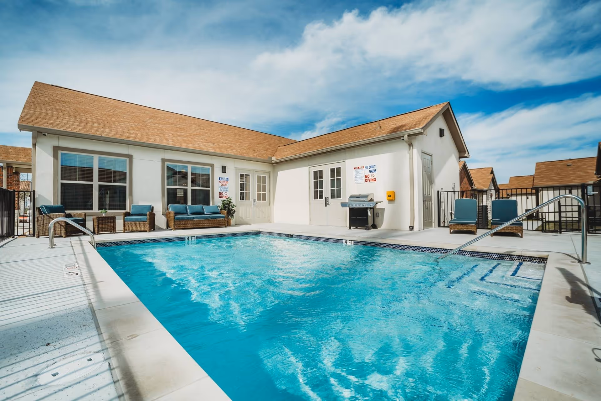 Outdoor swimming pool with clear blue water surrounded by a concrete deck. There are several cushioned wicker chairs and a sofa along the building wall. The building has white walls and a brown shingled roof. Signs on the wall indicate pool rules including 'No Diving'. A barbecue grill is also visible near the doors.