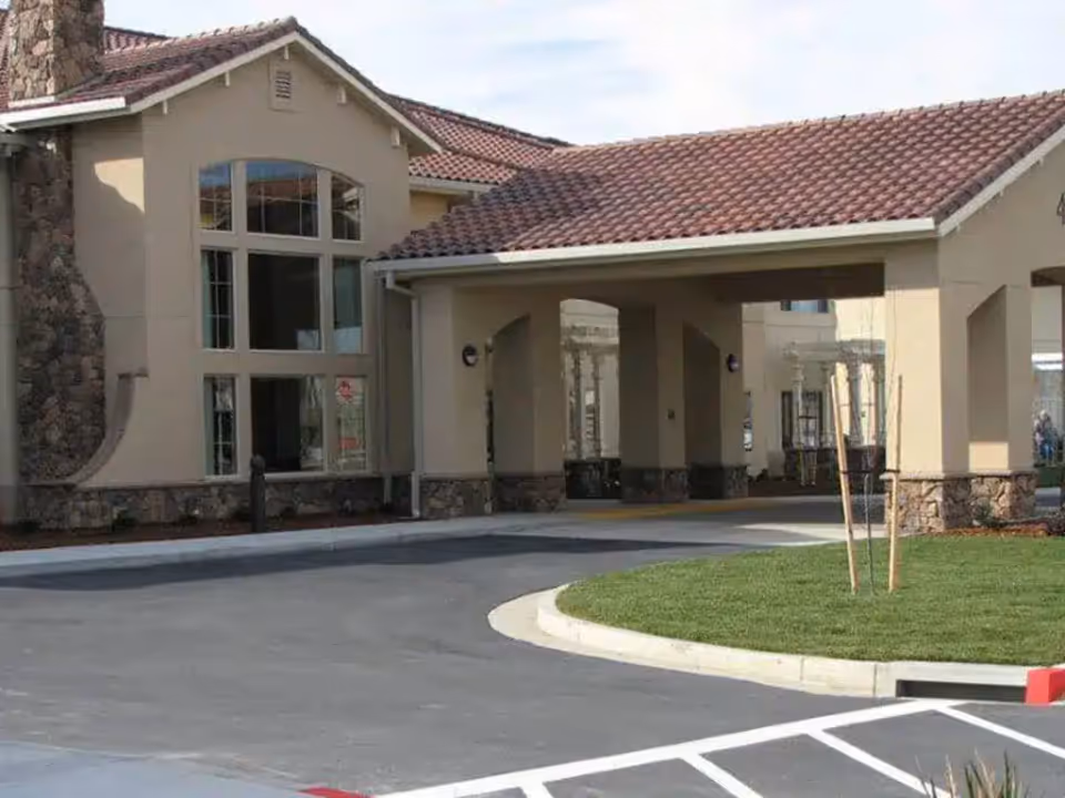 Covered driveway and entrance of a beige stucco building with a red tiled roof and large windows.