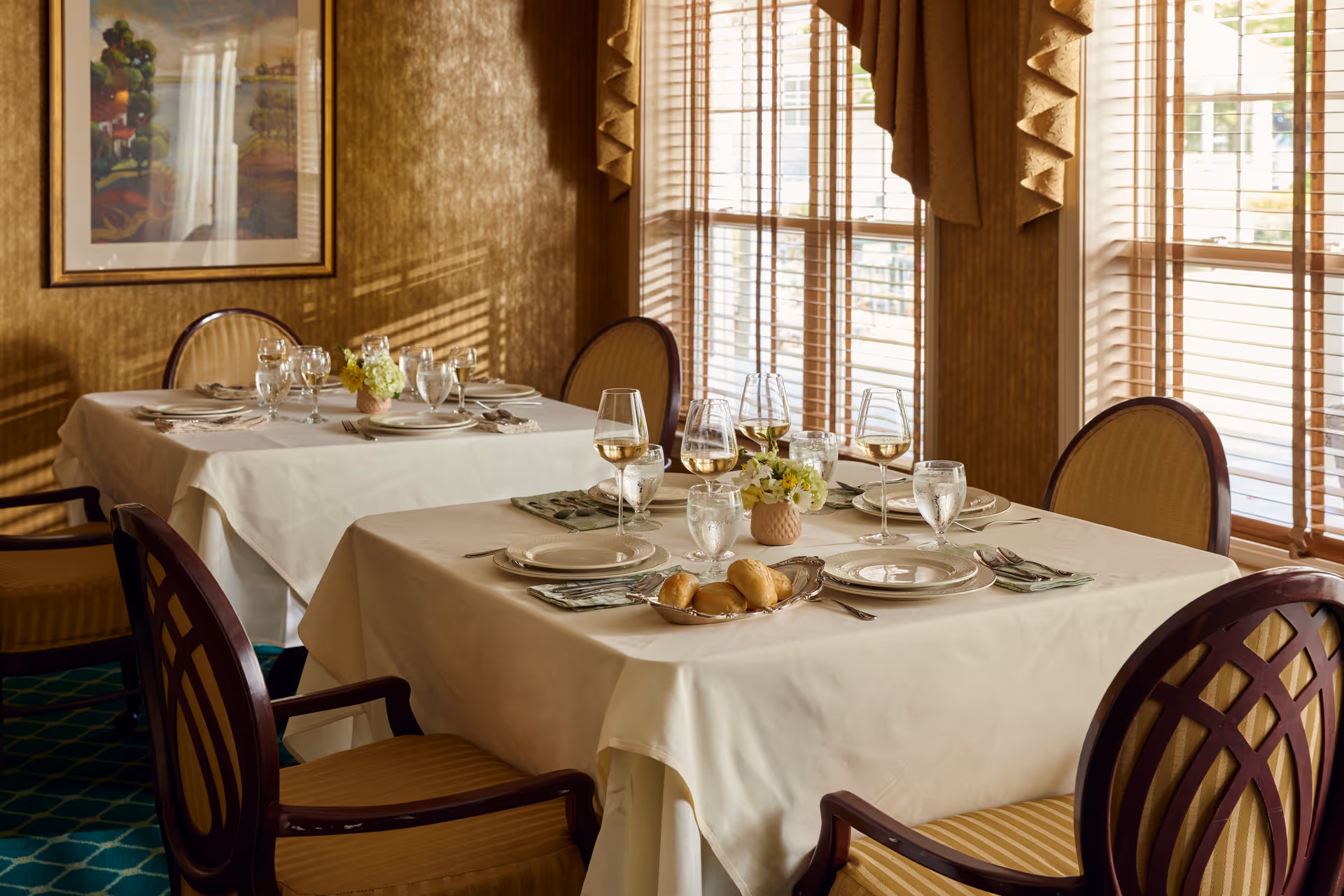 A dining room with two tables covered in white tablecloths, each set with plates, glasses of water and wine, silverware, cloth napkins, and small floral centerpieces. The room has large windows with wooden blinds allowing natural light to enter, and a framed painting on the wall.